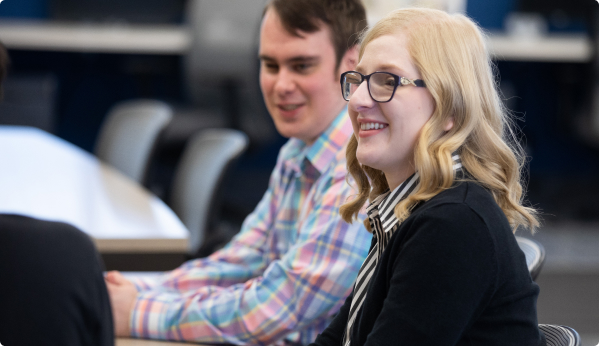 A young blonde woman wearing black glasses sites in a class room next to a brunette man in a colorful plaid shirt.