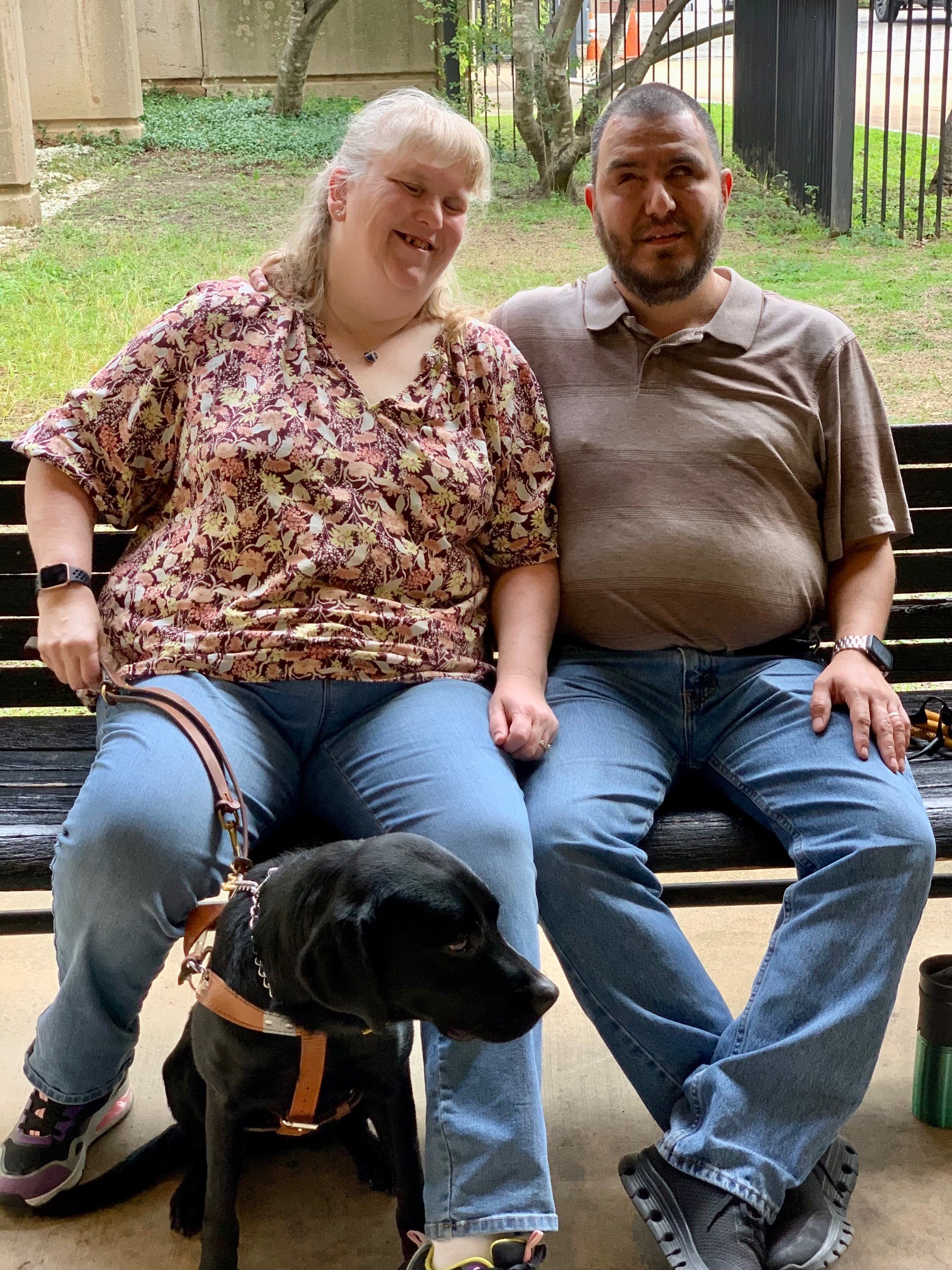 Keela and Ed sitting side-by-side on a park bench smiling with Keela's guide dog in between them