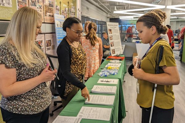 A young girl holding her white cane, talking to a professional at a career booth inside Envision.