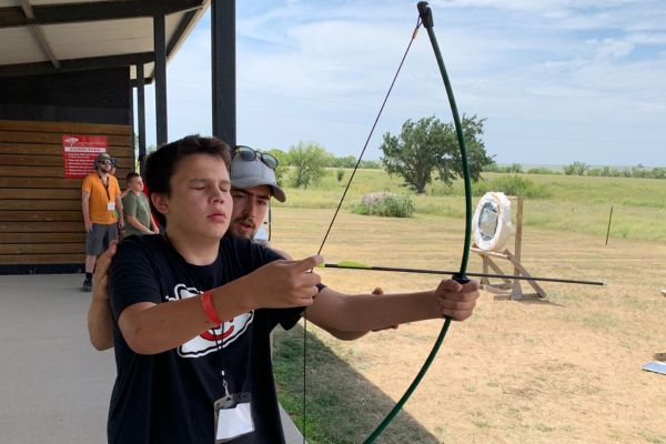 A counselor showing a visually impaired boy how to pull the bow of a bow and arrow.