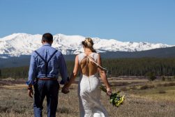 A bride and groom walking through a field with mountains in the background.